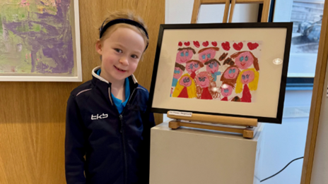 A young girl in a dark blue jacket smiling standing next to a child's painting of a group of people.
