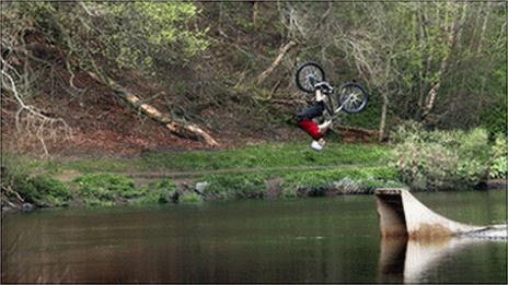 A mountain biker flips in the air froma ramp into the river.