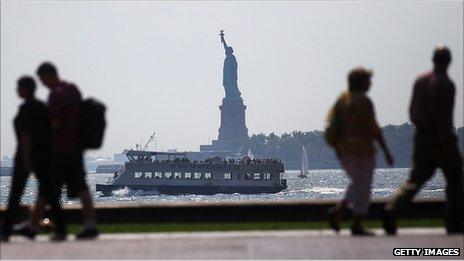 People walk along the waterfront in view of Liberty Island, New York