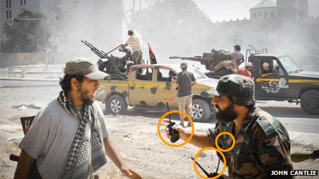 Scene in Sirte, with gun cars and two men talking in foreground - man on right with three radios