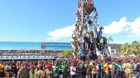 Thousands of Zambian people gather at Lusaka airport, some climbing on to a scaffold to get a glimpse of the football players