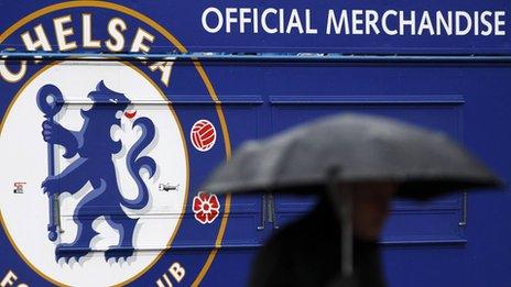 A man carrying an umbrella passes an closed merchandise kiosk at Chelsea football club.