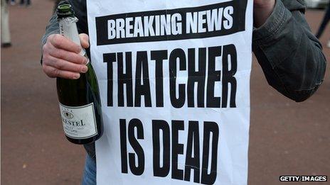 A man holds a bottle of champagne next to a newspaper poster carrying the news of Thatcher's death