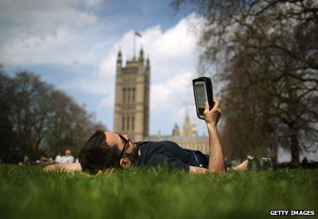 Man reads e-reader lying in grass near Houses of Parliament