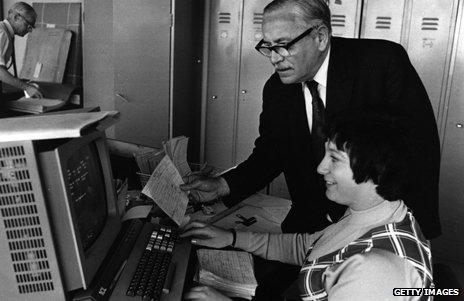 A man leans over the desk of a woman using one of the early word processors