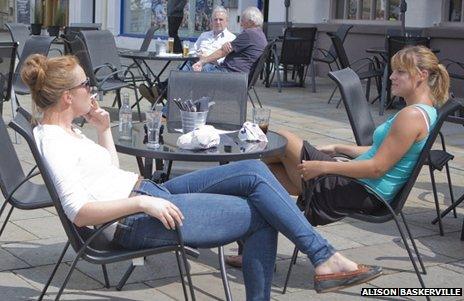 Two women sit outside a bar or pub, one is smoking