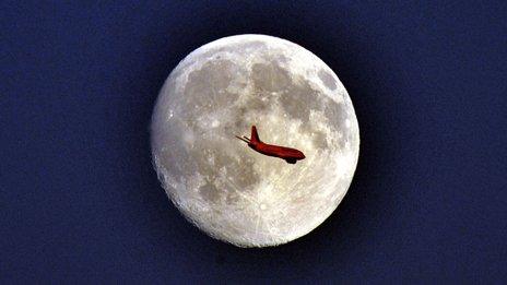 A plane crosses in front of a full moon. The plane is red from the setting sun