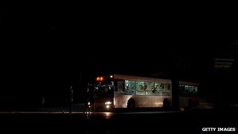 Cubans walk along a dark Havana street, lit only by bus headlights