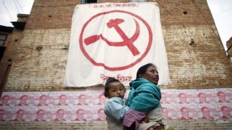 A Nepalese woman carries a child on her back while walking past a political flag and election campaign posters with portraits of Communist Party of Nepal (Maoist) Chairman Pushpa Kamal Dahal, in Kathmandu (31 October 2013)