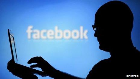 A man with a laptop is silhouetted against a screen with a Facebook logo in the background 14 August 2013