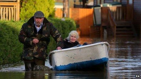 A man wades through water, pulling a boat in which a woman is sitting