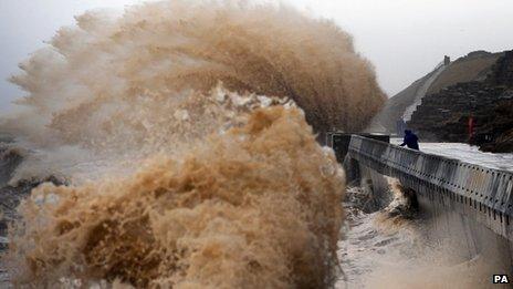 A huge wave breaks on a sea wall, behind which one person stands