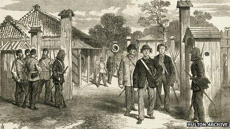 The entrance gate to the city of Yokohama, guarded by European troops at the time of the Meiji restoration, 1868. The three Japanese men walking through the gate are wearing Western style clothes.