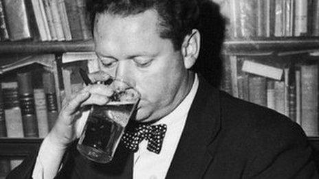 Welsh poet Dylan Thomas (1914 - 1953) drinking a glass of beer and smoking while seated at a desk with stacks of his books of poetry, New York City, c. 1950.