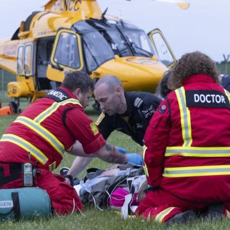 A doctor and a paramedic, dressed in red jackets with hi-vis stripes, and a pilot wearing a black shirt, kneel on a green field as they treat a patient with medical equipment. In the background is a yellow helicopter, its doors open. 