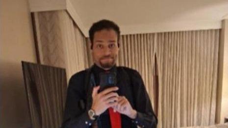 A man wearing a black button-down shirt and red tie holds a phone as he takes a photo of himself