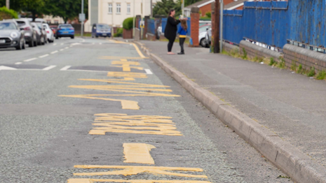 There are new road markings outside a school. Large yellow letters are painted on the road and several cars are parked on the left-hand side.