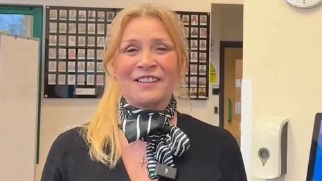 Cathy Hunt, a middle-aged woman with mid-length blonde hair, wearing a black neckerchief and a gold necklace with a cross pendant. She has a black blouse and stands in front of a grid showing small head and shoulders images of people.