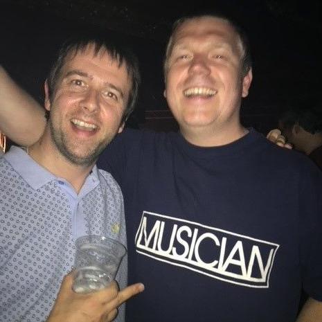 Mark Maloney and friend at a gig in the 90s. Mark is smiling and is holding a drink and pointing to his friend. His friend's shirt says "Musician".