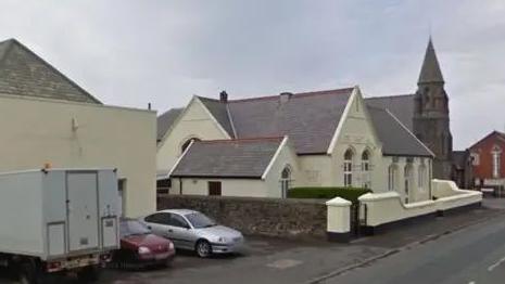 A street view of an old white chapel with the spire of a church visible behind it. There are cars parked to the left of the building.