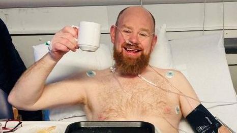 Man with ginger beard in hospital bed, with bare chest and hooked up to wires, smiling, and holding up a white mug by its handle in a 'cheers' gesture.