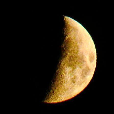 The moon, part illuminated against a black sky. It has a yellow tinge.