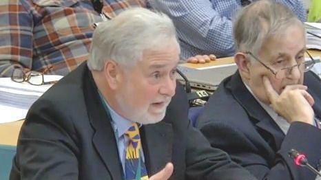 Councillor John Francis, who has grey hair and a grey beard, sits in a council chamber wearing a dark suit and a yellow, blue and green patterned tie. He is turned slightly away from the camera, looking towards a microphone during the meeting.