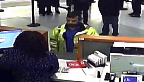 A man is seen in a CCTV screenshot. He is standing at a cashier's desk, wearing a yellow hi-vis jacket and a blue shirt. He has short black hair and a beard. We can see the back of the cashier's head, while others wait in line behind the man.