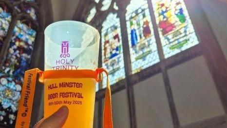 A beer cup is being held up against the backdrop of inside a church with a large stained-glass window with the sun peeking through. The cup says Holy Trinity Hull Minster Beer Festival on it with a yellow ribbon round it.