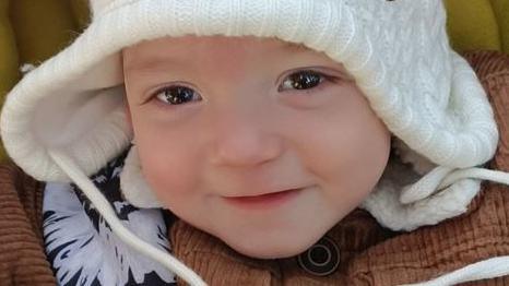 A young boy with large brown eyes smiles at the camera. He is wearing a white woolly hat