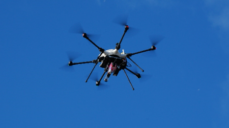 A grey and black drone in mid-flight. It has six "arms" and a red cylindrical device attached to the bottom of it.