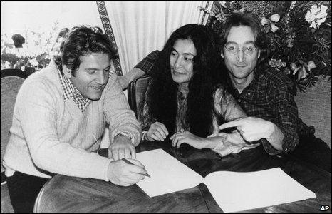 Allen Klein signs paperwork, with Yoko Ono and John Lennon sitting next to him at a small round table.  