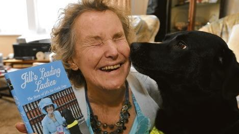 Jill Allen-King is smiling and holding a book. She is wearing a white and blue top with blue necklace and she is next to a large black dog.