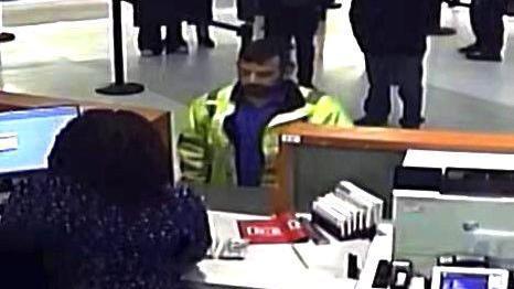 A CCTV screenshot of Matthew McDonagh standing at a cashier's desk, wearing a yellow hi-vis jacket and a blue shirt. He has short black hair and a beard. We can see the back of the cashier's head, while others wait in line behind the man.