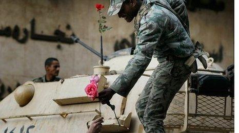 A smiling soldier on a tank being handed a rose by a protester