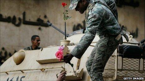 A smiling soldier on a tank being handed a rose by a protester