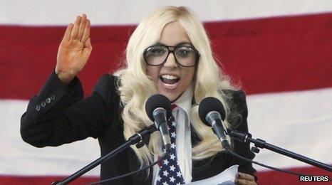 Lady Gaga dressed in blonde wig and wearing suit speaking at a rally