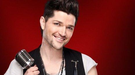 Danny O'Donoghue in his publicity photograph for 91ȱ The Voice. He is wearing a white t-shirt and a smart black waistcoat. His hair is coiffed into an impressive quiff! He is holding a retro microphone and smiling.