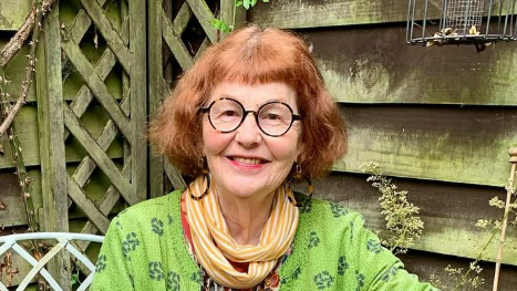 Marcia Riddington with short red hair and black glasses smiles at the camera while sitting in a garden in front of a shed. She is wearing a green top with a yellow and white striped scarf around her neck.