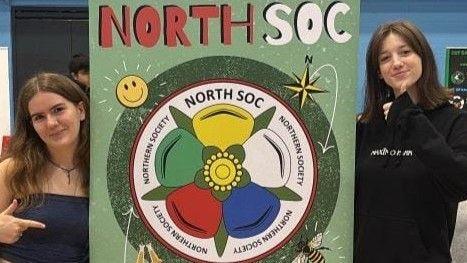 Two young women stand next to a banner promoting "North Soc" with a logo combining the Yorkshire and Lancashire roses.
