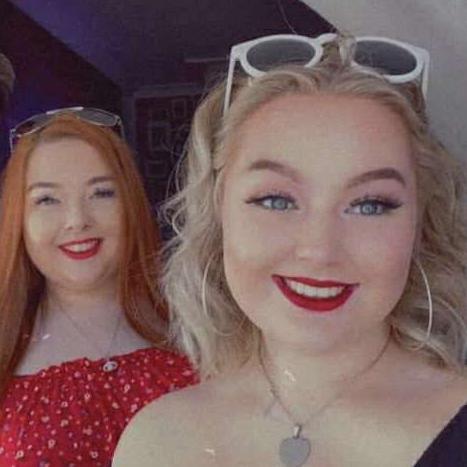 Two girls with sunglasses on their heads and red lipstick, smiling at the camera. One has long straight red hair, the other short curly blonde hair. The one with red hair is stood behind the other and has a red top on. They both silver have necklaces with pendants on. 