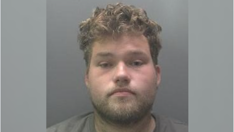 Luke Jarrett is in the middle of the image looking towards the camera. He has short brown hair and a beard and is wearing a grey T-shirt.