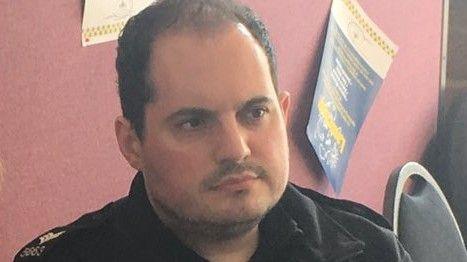 A close-up photograph of a police officer who has short dark curly hair. He is wearing a dark collared uniform and is seated against a pink wall with several posters or notices visible in the background. Part of a chair can be seen to the right-hand side of the image, while the lighting appears soft and natural. The image is cropped tightly around the subject, with the focus centred on the face and upper body.