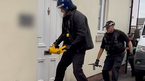 Three police officers approaching the front door of a flat. The first officer is about to break through the door using a yellow implement. He is wearing a helmet. 