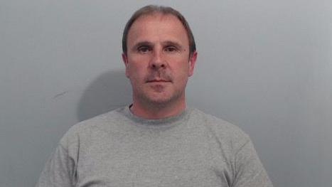 A mug shot of Irvine. It is a head and shoulders image, he is wearing a grey sweatshirt and has short brown hair. He looks directly at the camera. Behind him is a plain grey wall.
