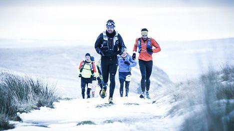 A group of runners appear on the brow of a hill. There is snow on the ground and they are all wrapped up in warm running gear.  