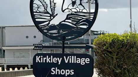 A black sign reading "Kirkley Village Shops" with an arrow pointing to shopping area can be seen in a seafront setting