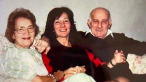 Jeanette sits between her parents on a sofa, all of them with their arms around each other. She has dark hair and is wearing a black top or jumper. Mascha is sat on her left, wearing a pale green patterned shirt, a bracelet and metallic-framed glasses. Jeanette's father is sat to the right, wearing a dark jumper and a white collared shirt. 