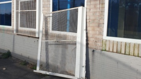 A large white meshed metal shutter is leant against the wall of a building