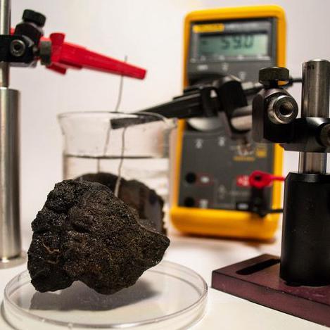 A metal nodule collected from the deep sea is probed with a voltmeter on a lab bench